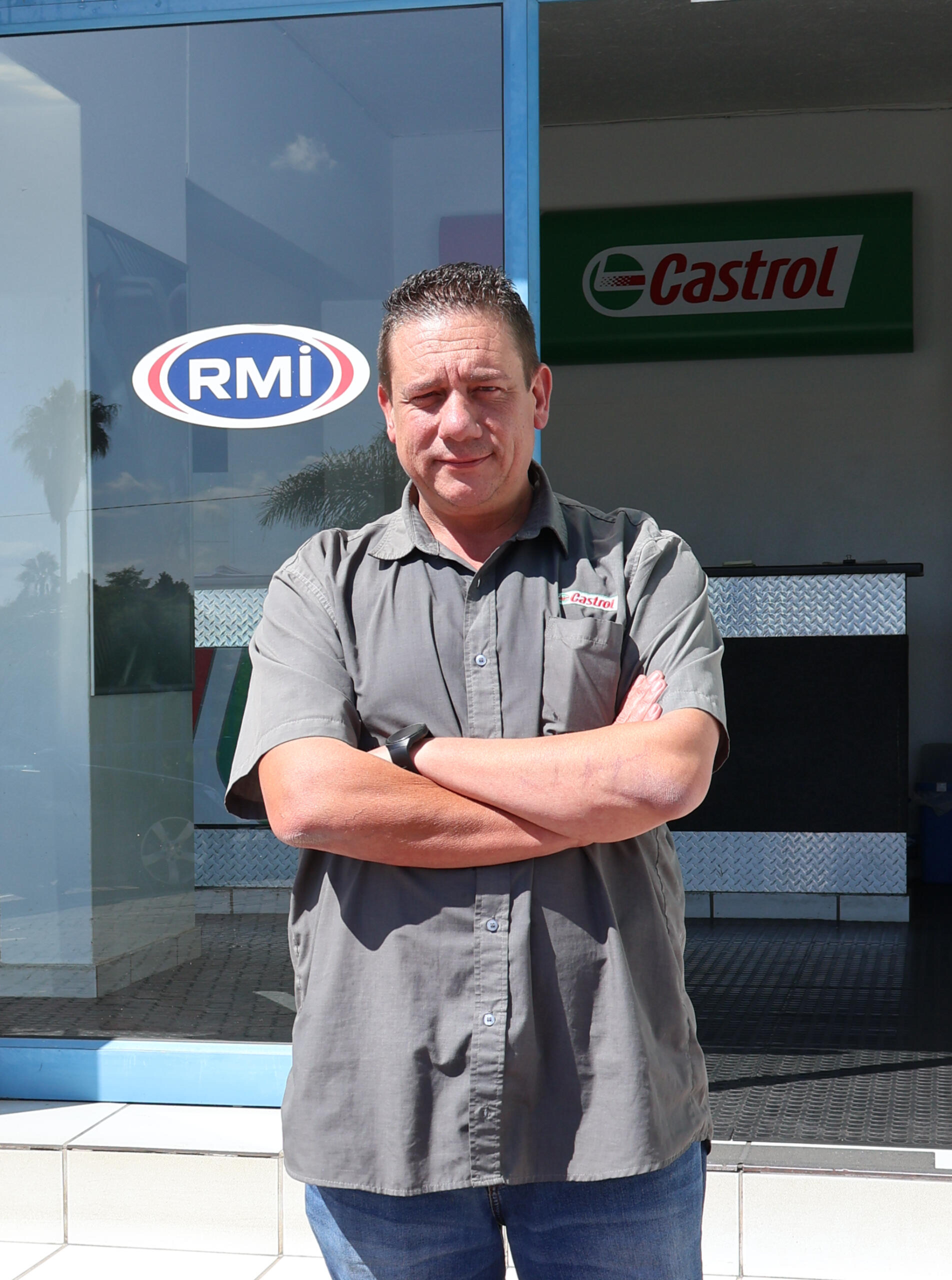 Manager Casper, standing with arms folded, wearing Castrol branded grey shirt, standing outside of the Castrol Auto Service Northcliff building. RMI logo in the background, on the glass door of the building. Castrol logo also in background.